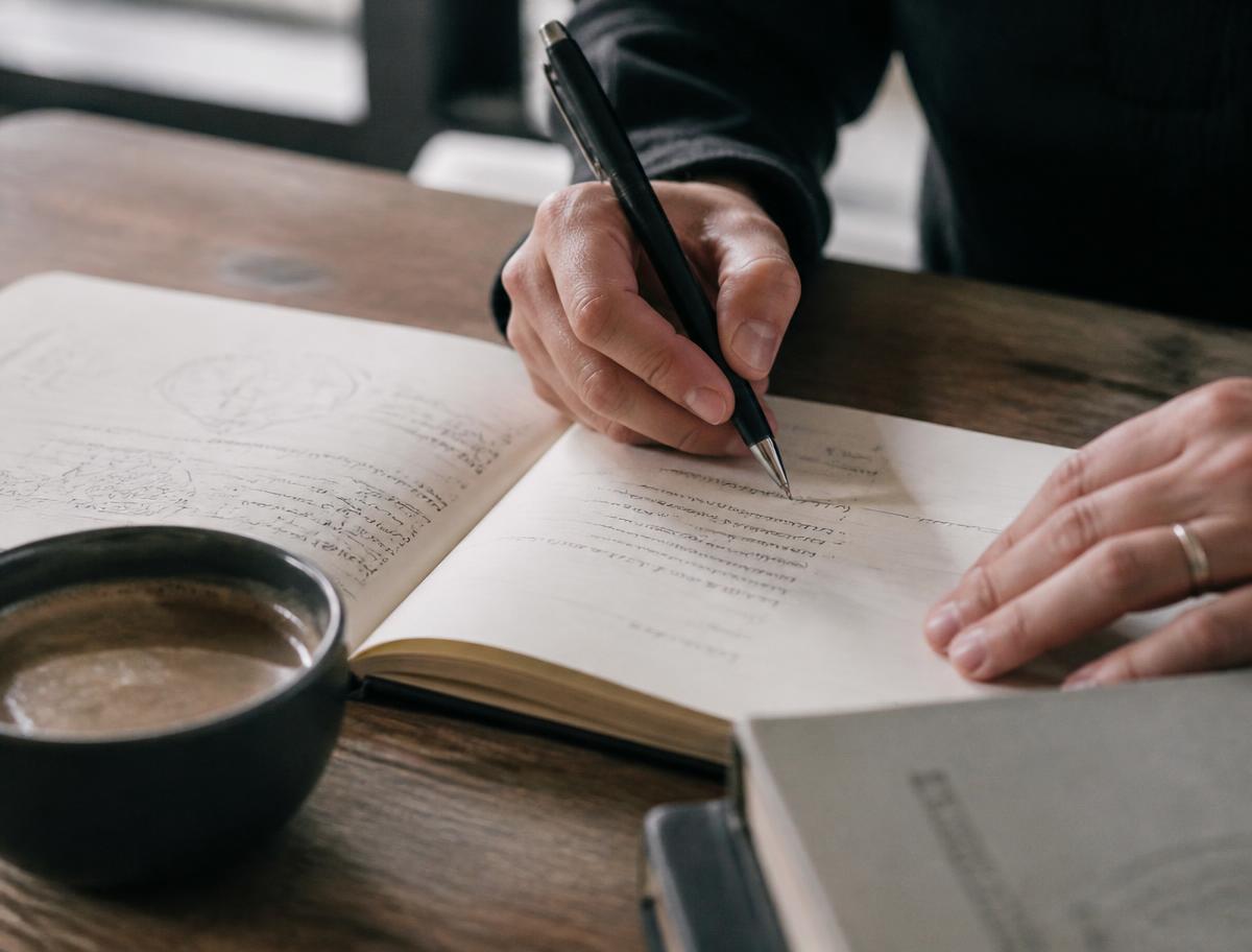 Mains en train d'écrire des notes au stylo dans un carnet, à côté d'une tasse de café